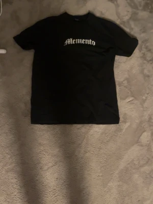 Svart Memento t-shirt med tryck - Säljer en svart t-shirt med texten 'Memento' i vit gotisk stil på bröstet och ett stort, detaljerat tryck på ryggen med text och grafiska element. T-shirten har klassisk passform och är kortärmad, perfekt för dig som gillar streetwear vibes.