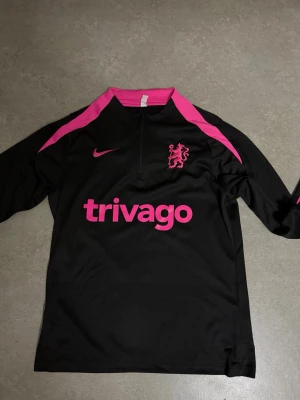 Svart Nike träningsjacka med rosa detaljer - Snygg svart träningsjacka från Nike med rosa detaljer och halv dragkedja. Jackan har rosa logga, klubbmärke och trivago-tryck på bröstet samt rosa paneler längs axlar och krage. Perfekt för träning eller sportiga outfits.