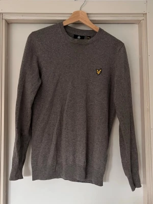 Grå stickad tröja från Lyle & Scott - Säljer en stilren grå stickad tröja från Lyle & Scott med deras klassiska gula fågel-logga på bröstet. Tröjan har rund halsringning, långa ärmar och ribbade muddar vid ärmslut och nederkant. Perfekt till lager på lager och enkel att matcha. Tröjan är i storlek XS