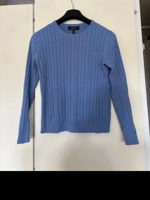 Blå kabelstickad tröja från GANT - Snygg ljusblå kabelstickad tröja från GANT med rund hals och diskret logotyp broderad på bröstet. Tröjan har långa ärmar och ribbade muddar. Perfekt för dig som gillar klassisk och stilren design. Materialet är mjukt och bekvämt.