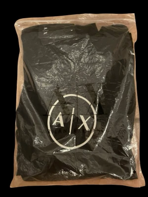 Svart ny t-shirt från Armani Exchange - Snygg svart t-shirt från Armani Exchange med stort vitt A|X-logotryck på bröstet. Klassisk passform och rund halsringning. Perfekt till jeans eller shorts för en clean och modern look. 