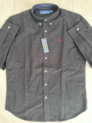 Svart skjorta från Ralph Lauren - Klassisk svart skjorta från Ralph Lauren med korta ärmar och knappdetaljer. Skjortan har en röd broderad logga på bröstet och button-down krage. Tillverkad i bomull för en skön känsla och stilren look.