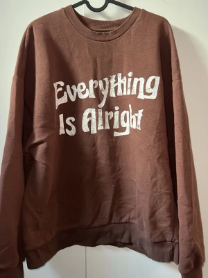 Brun sweatshirt från Gina Tricot - Säljer en brun sweatshirt från Gina Tricot med texten 'Everything Is Alright' i vitt framtill. Texten är lite urtvättad. 