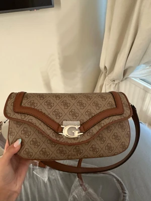 Beige axelväska från Guess - Snygg beige axelväska från Guess med brunt mönster och detaljer i brunt konstläder. Väskan har ett stort G-spänne i guld framtill och magnetlås. Invändigt finns flera fack för bra organisering. Perfekt för dig som gillar stilrena accessoarer.