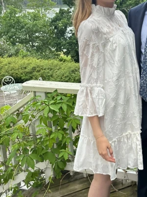 Vit blommig klänning med volang - Romantisk vit klänning med hög krage, blommigt mönster och volangdetaljer på ärmar och nederkant. Klänningen har trekvartsärm och är tillverkad i ett lätt, luftigt material som passar perfekt till sommarens tillfällen.
