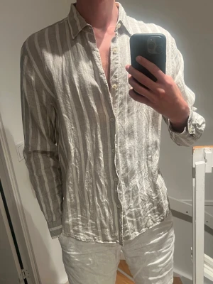 Randig linneskjorta i beige och vit - Säljer en snygg randig skjorta i beige och vitt med klassisk krage och knappar framtill. Skjortan är tillverkad i linne vilket gör den luftig och perfekt för varma dagar. Långärmad modell med relaxed fit som ger en chill vibe.