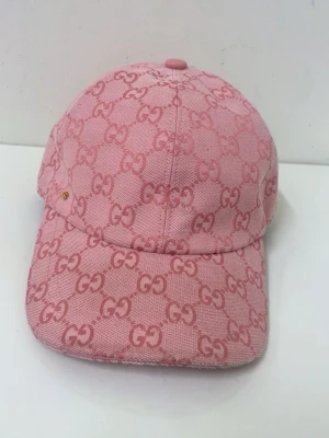 Rosa keps från Gucci med GG-mönster - Säljer en snygg rosa keps från Gucci med det klassiska GG-monogrammet över hela kepsen. Kepsen har böjd skärm och justerbart band bak. Tillverkad i tyg med exklusiv känsla och detaljer i rosa. Perfekt för dig som vill sticka ut med stil.