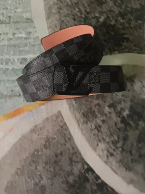 Svart rutigt L V-bälte i skinn - Snyggt svart bälte från L V med klassiskt rutigt mönster och L V-logga i metall på spännet. Bältet är gjort i skinn och har en stilren design som passar till många olika outfits.
