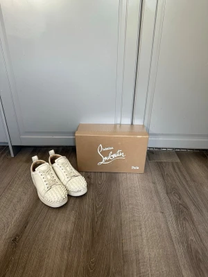 Vita Christian Louboutin  - Säljer ett par vita sneakers från Christian Louboutin med ikoniska röda sulor och coola nitar över hela tån. Skorna har lågt skaft, snörning och är tillverkade i läder. Perfekta för dig som vill sticka ut med en lyxig och edgy stil. Hör av för frågor och funderingar 