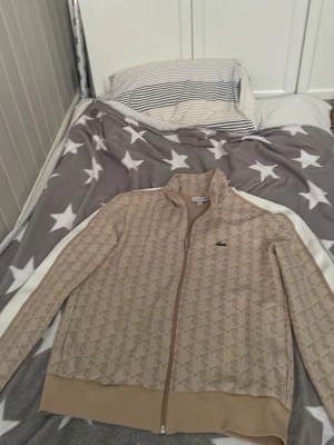 Beige ziptröja från Lacoste - Snygg beige ziptröja från Lacoste med diskret mönster och klassisk grön krokodillogga på bröstet. Tröjan har hög krage, hel dragkedja och ribbade muddar. Perfekt för en sportig och trendig look.