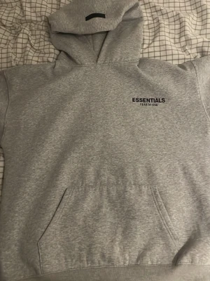 Grå Essentials Fear of God hoodie - Snygg grå hoodie från Essentials Fear of God med svart logga på bröstet. Klassisk känguruficka framtill och stor huva. Tillverkad i mjukt bomullsmaterial som känns riktigt skönt mot huden. Perfekt för dig som gillar streetwear och enkel stil.