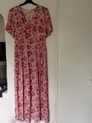 Blommig rosa långklänning  - Lång klänning i rosa med blommigt mönster i rött och rosa. Klänningen har korta ärmar, markerad midja med ett band att knyta i midjan och ett luftigt fall. Perfekt för sommarens alla tillfällen. Materialet är mjukt och svalt, troligen bomull eller polyester. Använd 1 gång. Köpt på MQ. Pris går att diskutera.