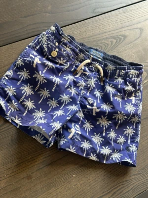 Blå badbyxor från Polo Ralph Lauren - Snygga blå badbyxor från Polo Ralph Lauren med vitt palm- och björnmönster. Klassisk modell med resår i midjan, snörning och ficka med knapp bak. Perfekta för stranden eller poolen. Materialet är snabbtorkande syntet. Storlek 5 år 