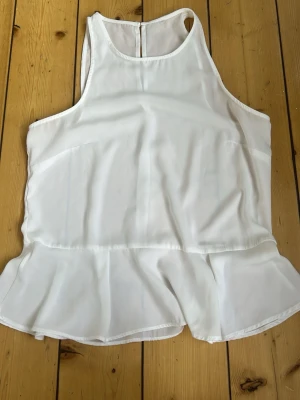 Vit volangtop med ärmlös design - Snygg vit topp med volangkant nedtill och ärmlös modell. Toppen har rund halsringning och är tillverkad i ett tunt, lätt material som ger en luftig känsla. Perfekt för varma dagar och enkel att styla med jeans eller kjol.