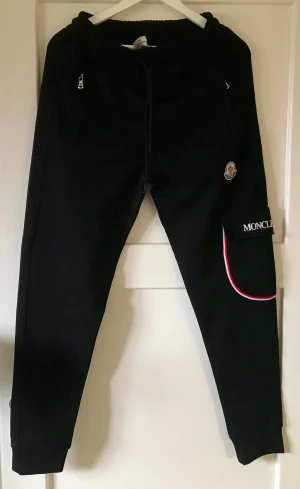 Svarta mjukisbyxor från Moncler - Snygga svarta mjukisbyxor från Moncler med dragsko i midjan, två fickor med dragkedja och en extra ficka på benet med Moncler-logga och rödvit detalj. Perfekta för chill eller streetwear. Sköna och stilrena med elastiska benslut.