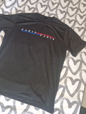 Tröja - Snygg svart t-shirt med texten 'PARIS PARIS' framtill och två linjer i rött och blått. Klassisk rund hals och korta ärmar. Perfekt för dig som gillar enkel men stilren design. 