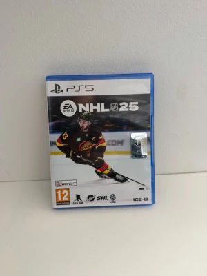NHL 25 PS5 spel - NHL 25 till PlayStation 5, ett ishockeyspel från EA Sports. Omslaget har en spelare i svart och gult på isen. Blå plastfodral med spelmanual och skiva med färgglad grafik. Perfekt för dig som gillar sportspel och hockey. Använd men i gott skick