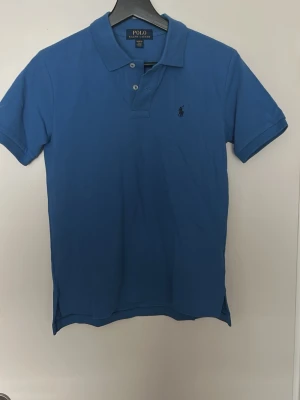 Blå pikétröja från Polo Ralph Lauren - Klassisk blå pikétröja från Polo Ralph Lauren med korta ärmar och krage. Storlek M(10-12) sitter som herr xs.