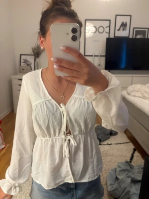 Vit blus med knytning framtill - Säljer en söt vit blus med långa ärmar och knytning framtill. Blusen har en djup v-ringning och lätt puffiga ärmar. Materialet är tunt och luftigt, perfekt för en trendig och avslappnad look. Passar till både jeans och kjol. Passar säkert s också 