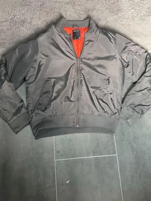 Grå bomberjacka med orange foder - Snygg grå bomberjacka med klassiskt orange foder. Jackan har ribbade muddar vid ärmslut och midja, två snedställda fickor fram och en ärmficka med dragkedja. Tillverkad i glansigt syntetmaterial för en modern look.