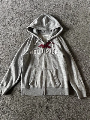 Hollister zip-hoodie - Snygg grå hoodie från Hollister med dragkedja, huva med vita snören och broderad logga i vitt och vinrött framtill. Mjuk bomullsblandning och två fickor fram. Perfekt för en avslappnad och cool stil.