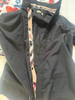 Svart Burberry skjorta med rutiga detaljer - Snygg svart skjorta från Burberry i storlek M. Klassisk modell med knappar framtill och ikoniska Burberry-rutiga detaljer längs knappslån. Tillverkad i mjuk bomull som känns skön mot huden. Perfekt för dig som vill ha en stilren look med exklusiv känsla. Passar M-S finns i Göteborg 