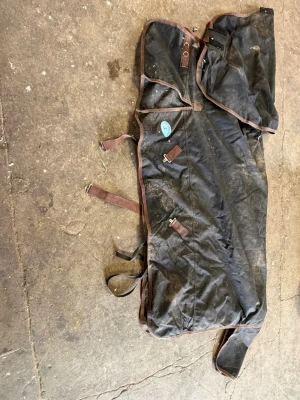 Svart hästtäcke med bruna detaljer - Säljer ett svart hästtäcke med bruna kantband och spännen. Täcket har flera remmar och spännen för att fästa runt hästen samt en hals.  Materialet är slitstarkt tyg och det finns en blå etikett på sidan ”tyfon”. Vintertäcke 400g endast använt denna vintern. Storlek 145cm. Helt men smutsigt. 