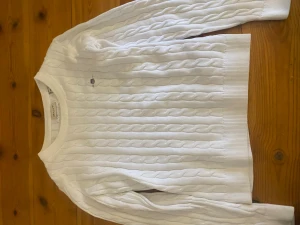 Vit kabelstickad tröja Gant - Kabelstickad vit tröja från Gant med rund halsringning och broderad logotyp på bröstet. Tröjan har ribbade muddar vid ärmslut och nederkant, och är tillverkad i mjukt material som ger en klassisk och stilren look.