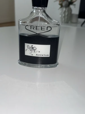 Creed Aventus Eau de Parfum 100ml - Creed Aventus en lyxig parfym i en elegant, genomskinlig glasflaska med svart etikett och silverdetaljer. Flaskan har en stilren form och pryds av en ikonisk ryttare. En klassiker från Creed som passar dig som gillar exklusiva dofter. Det enda problemet är att något har fastnat vid sprayaren så att korken inte sitter helt fast men det påverkar inte hur den sprayar den sprayar fortfarande som den ska. Jag uppskattar ungefär 93 ml av 100 ml. Jag säljer den då den inte riktigt passar mig. 100% äkta