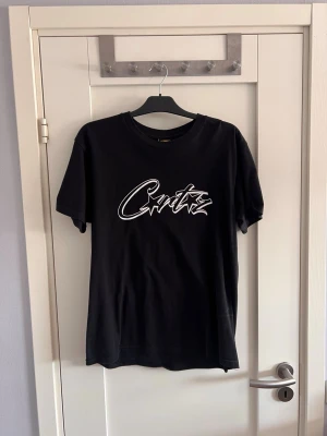 Svart t-shirt från Corteiz - Säljer en svart t-shirt från Crtz med vit grafisk logga på bröstet. Klassisk rund halsringning och korta ärmar. Tillverkad i mjuk bomull som känns skön mot huden. Perfekt för en avslappnad och stilren look.