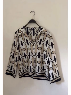 Mönstrad blus från Zara - Snygg satinblus från Zara med svart och beige grafiskt mönster på vit botten. Blusen har krage, knäppning framtill och långa ärmar. Tillverkad i ett lätt och luftigt material som ger en avslappnad men trendig look.
