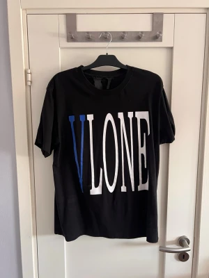 Svart VLONE t-shirt med tryck - Säljer en svart t-shirt från VLONE med stort vitt och blått VLONE-tryck på framsidan. Baksidan har ett blått V med en vit orm runt. Klassisk rund hals och korta ärmar. Perfekt för dig som gillar streetwear och vill sticka ut.