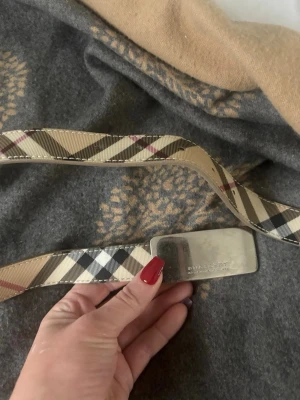 Beige rutigt bälte från Burberry - Snyggt bälte från Burberry med klassiskt beige, svart och rött rutmönster. Bältet har en silverfärgad metallspänne med Burberry-logga. Perfekt accessoar för att lyfta vilken outfit som helst.
