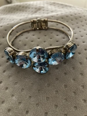 Silverfärgat armband med blå stenar - Snyggt armband i silverfärgad metall med stora, runda blå glittrande stenar på rad. Armbandet har ett smidigt gångjärn och ger en riktigt statement-look. Perfekt för dig som vill sticka ut med accessoarerna och gillar bling.