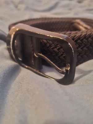Mörkbrunt flätat bälte - Snyggt mörkbrunt flätat bälte med metallspänne. Bältet har en stilren design och passar perfekt till jeans eller chinos. Flätningen ger en cool struktur och gör det enkelt att justera längden. Perfekt accessoar för att lyfta din outfit.