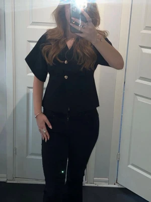 Svart blus med knappar - Snygg svart blus med v-ringning och korta vida ärmar. Blusen har två dekorativa guldknappar framtill och en rak passform som ger en stilren look. Perfekt att styla med jeans eller kjol för en trendig outfit.
