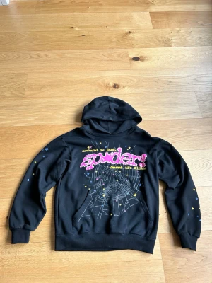 Svart hoodie från Spider - Svart hoodie från Spider Worldwide med stor färgglad grafisk text och spindelnätsmönster på ryggen. Hoodien har huva, magficka och små stjärnor på ärmarna. Materialet är mjukt och bekvämt, Har använts 4 gånger.