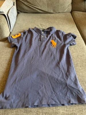 Blå pikétröja från Polo Ralph Lauren - Säljer en blå pikétröja från Polo Ralph Lauren med orange logga på bröstet och siffran 3 på ärmen. Klassisk krage med knappar och korta ärmar. Modellen är custom fit och materialet är bomull. Perfekt för en avslappnad men snygg stil.