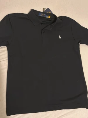 Svart pikétröja från Polo Ralph Lauren - Klassisk svart pikétröja från Polo Ralph Lauren med vit broderad logga på bröstet. Tröjan har krage, knappar framtill och är tillverkad i bomull för en bekväm känsla. Perfekt till jeans eller shorts för en clean look.
