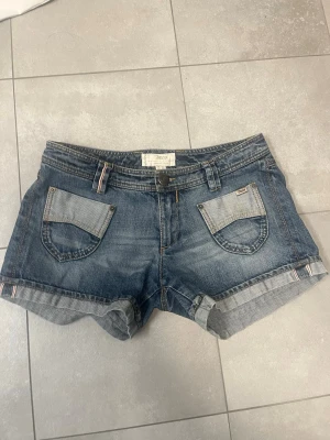 Blå lågmidjade korta  jeansshorts - Snygga blå jeansshorts med uppvikta ben och två framfickor med dekorativa detaljer. Klassisk bälteshällor. Korta och lågmidjade- precis vad som trendar