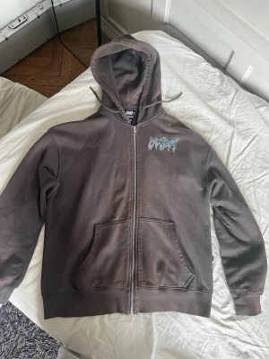 Mörkgrå zip hoodie från Sweet SKTBS - Säljer en mörkgrå hoodie från Sweet sktbs med dragkedja, stor huva och fickor framtill. Tryck i ljusblått och vitt på bröstet och stort grafiskt tryck på ryggen. Perfekt för dig som gillar streetwear.