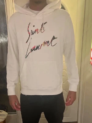 Vit Saint Laurent hoodie med tryck - Snygg vit hoodie från Saint Laurent med färgglatt logotyp-tryck på bröstet. Hoodien har  magficka och ribbade muddar vid ärmslut och nederkant. Perfekt för en clean och trendig look. Modellen är 180 cm och väger ca 65 kg