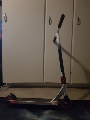 NKD trick sparkcykel silver/svart - Säljer en stilren trick sparkcykel från NKD i silver och svart med röda detaljer. Handtagen är röda och däcket har ett mönstrat grepp. Perfekt för dig som gillar att åka i skateparken eller bara vill cruisa runt nu till sommaren 