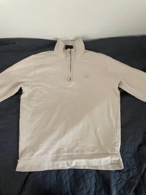 Beige halfzip från Lyle & Scott - Snygg beige halfzip-tröja från Lyle & Scott med diskret broderad logga på bröstet. Tröjan har hög krage, dragkedja framtill och är tillverkad i mjukt bomullsmaterial. Perfekt för en clean och stilren look.