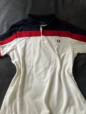 Vit och marinblå piké från Fred Perry - Säljer en snygg pikétröja från Fred Perry i vitt med marinblå och röd panel upptill. Tröjan har klassisk krage, knappar vid halsen och den ikoniska Fred Perry-loggan broderad på bröstet. Perfekt till jeans eller shorts för en sportig look.