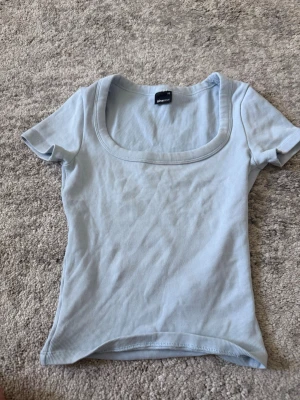 Ljusblå ribbad topp från Gina Tricot - Supersöt ljusblå ribbad topp från Gina Tricot med korta ärmar och rundad, djup urringning. Perfekt att styla med jeans eller kjol för en fräsch look. Materialet är mjukt och stretchigt för en bekväm passform.