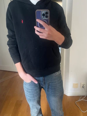 Svart hoodie från Polo Ralph Lauren - Klassisk svart hoodie från Polo Ralph Lauren med röd broderad logga på bröstet. Hoodien har huva, känguruficka och ribbade muddar. Tillverkad i mjukt bomullsmaterial som är skönt att bära. Perfekt för en avslappnad och stilren look.