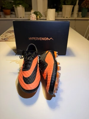 Nike Hypervenom orange/svarta fotbollsskor - Säljer ett par Nike Hypervenom fotbollsskor i orange och svart med klassisk Nike-logga på sidan. Skorna har en lätt och slitstark ovandel i syntetmaterial och fasta dobbar för spel på gräs. Snygg, modern design som sticker ut på planen. ALLT MEDFÖLJER!!! Hör av er vid frågor! 
