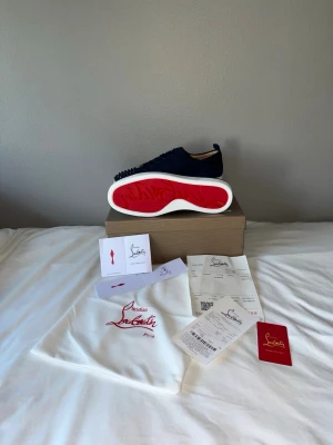 Christian Louboutin sneakers - Snygga marinblå sneakers från Christian Louboutin med klassisk röd sula och coola svarta nitar på tån. Skorna är låga och har ovandel i mocka, vita sulor och matchande marinblå skosnören. Perfekta för dig som vill sticka ut med exklusiv design.