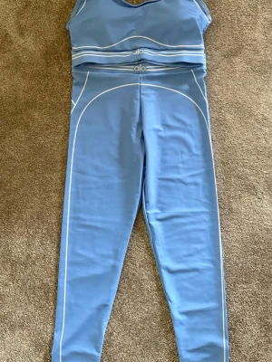 Blått träningsset från Alo Yoga - Snyggt blått träningsset från Alo Yoga med vita detaljer. Setet består av en croppad topp med breda axelband och matchande tights med hög midja. Materialet är stretchigt och passar perfekt för träning. Diskreta loggor på både topp och byxa.